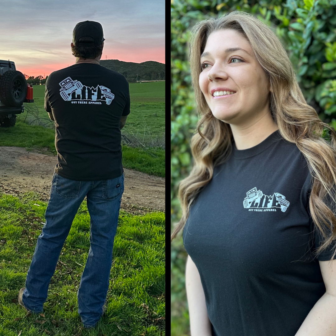 A man and a woman wearing black Life Out There Apparel's Unisex The Crawler T Shirts embody the essence of off-roading adventure. The man faces away, likely dreaming of the excitement of rock crawling ahead, while the woman smiles brightly next to him, surrounded by a lush green backdrop.
