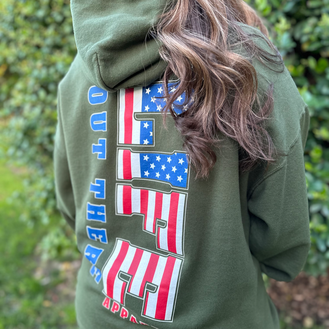 A person with long hair is wearing a military green "Unisex The Freedom Hoodie" from Life Out There Apparel, showcasing a design on the back featuring the American flag and the text "Out There Apparel." Embracing THE FREEDOM, they are outdoors, standing amidst greenery.