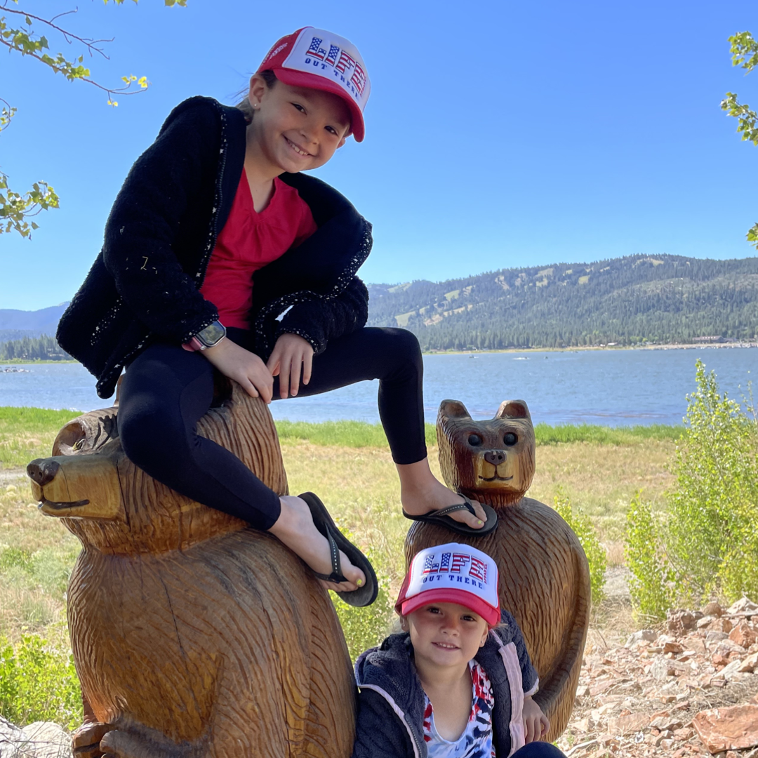 Two children in matching red and white Youth The Freedom Embroidered Snapback Trucker Hats from Life Out There Apparel sit and stand on carved wooden bear statues. They are outdoors near a lake, surrounded by green hills and trees under a clear blue sky, embodying the freedom and spirit of the 4th of July.