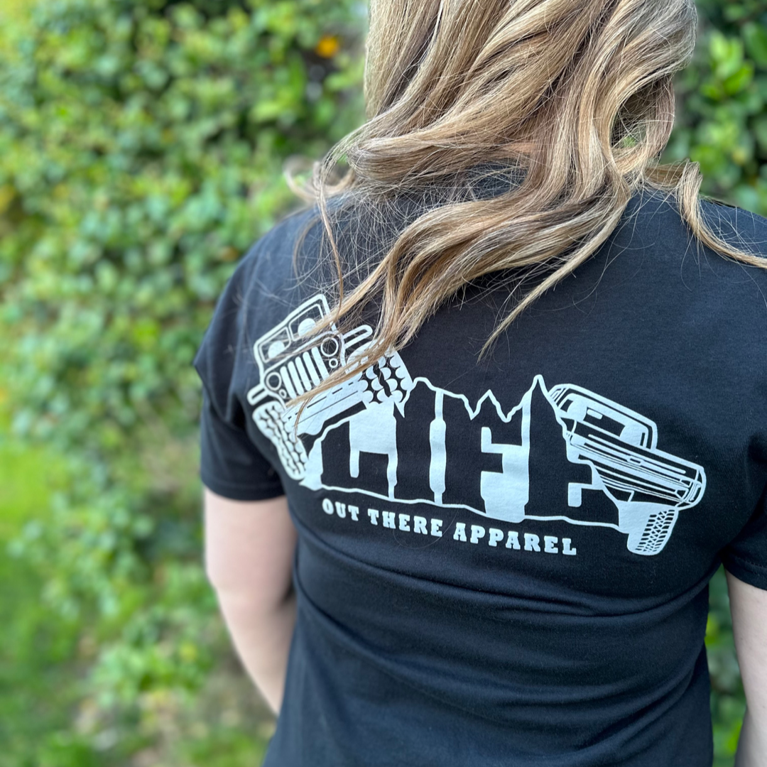 A person with wavy hair stands outdoors, wearing the Unisex The Crawler T Shirt - Black from Life Out There Apparel, featuring designs of a vehicle and trees along with the word LIFE. The text "Life Out There Apparel" below suggests off-roading adventures, all surrounded by greenery.