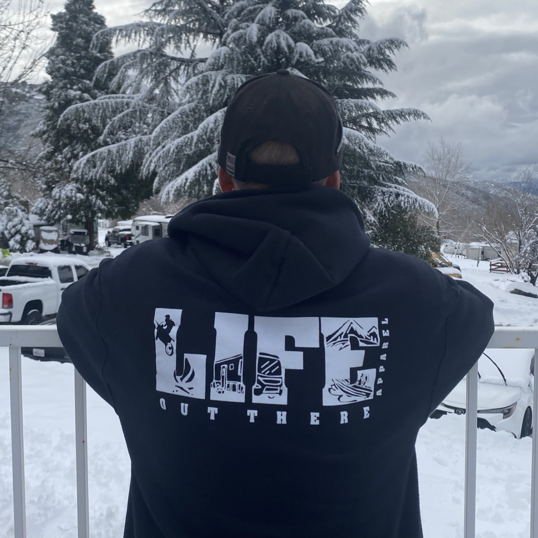 A person wearing the Unisex The OG Hoodie in Black/White by Life Out There Apparel stands on a snowy deck, prepared for a day of snowboarding. Pine trees and snow-covered vehicles are visible in the background beneath an overcast sky.