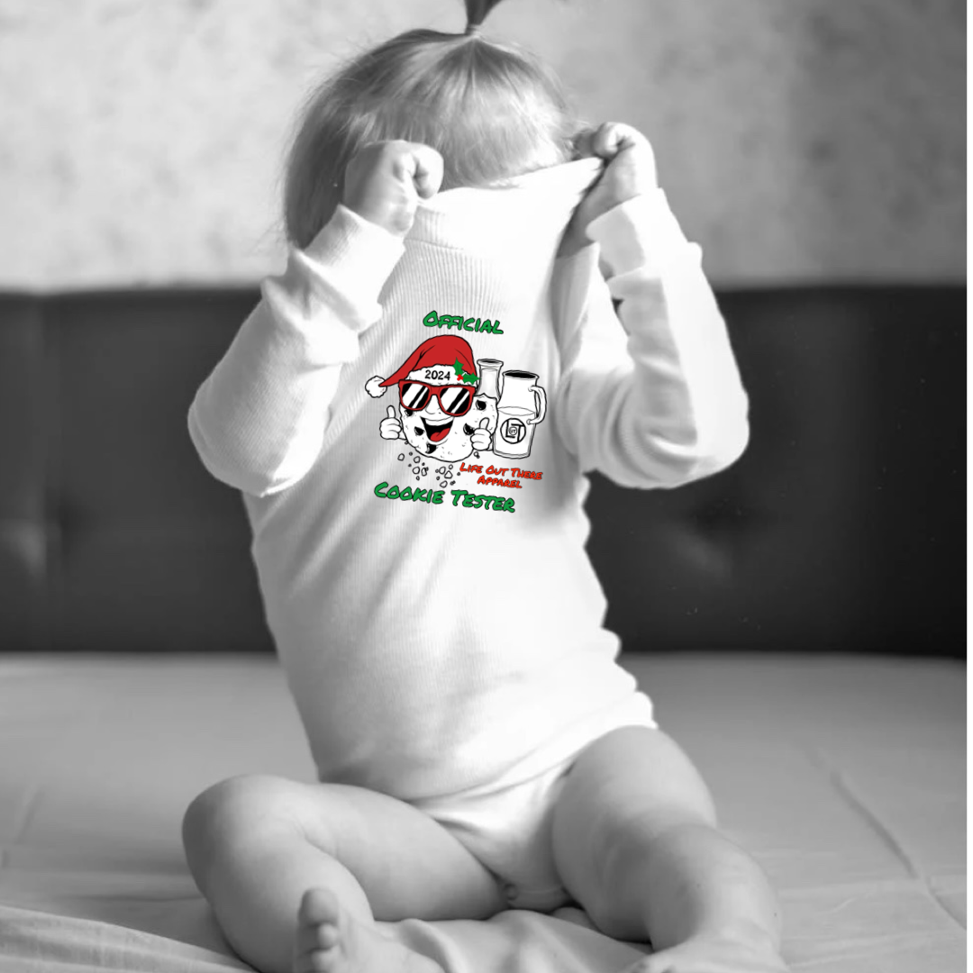 White long sleeve baby shirt with a cartoon cookie wearing a Santa hat and sunglasses labeled "2024," alongside glass milk cartons. Text reads, "Life Out The Apparel / official cookie tester”.