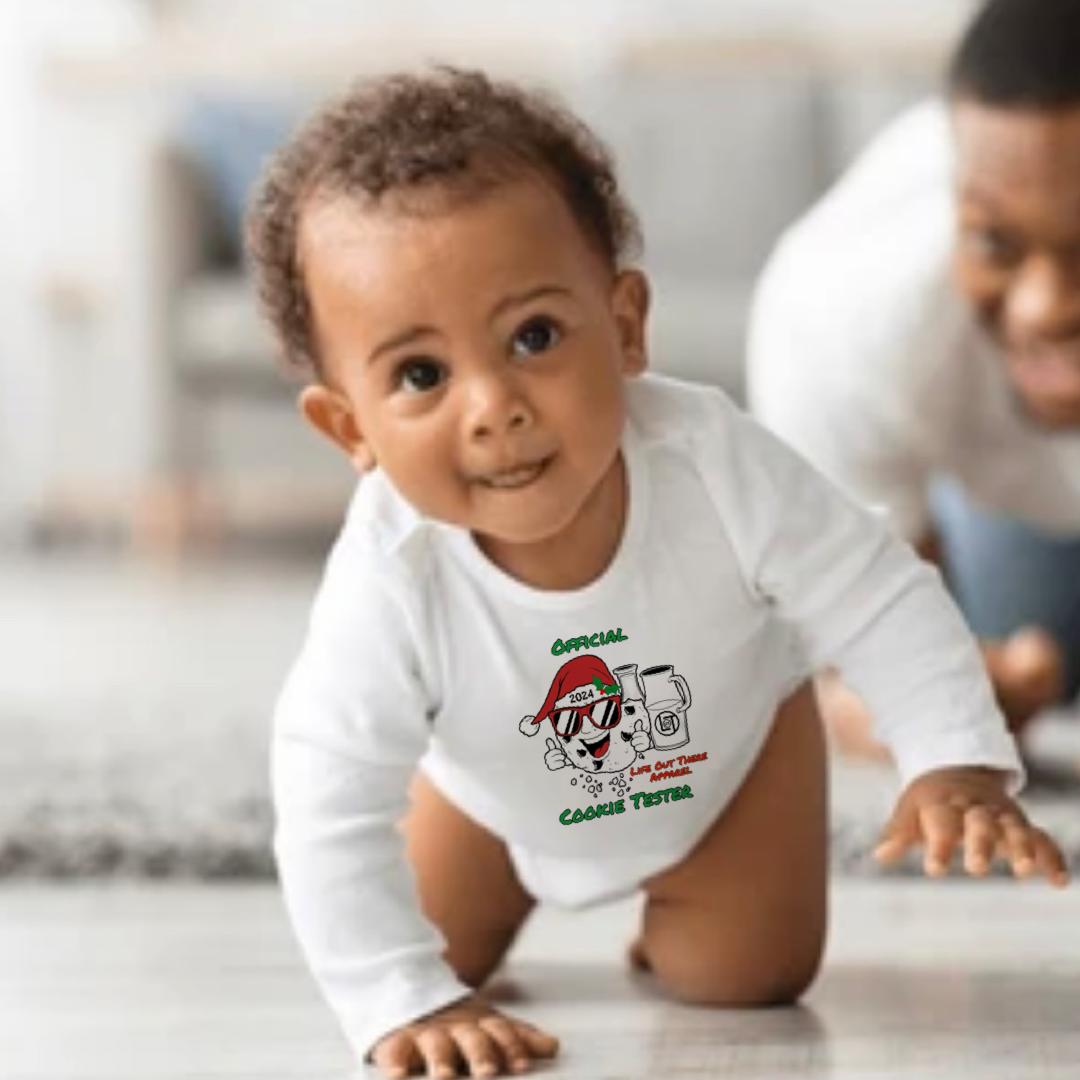 White long sleeve baby shirt with a cartoon cookie wearing a Santa hat and sunglasses labeled "2024," alongside glass milk cartons. Text reads, "Life Out The Apparel / official cookie tester”.