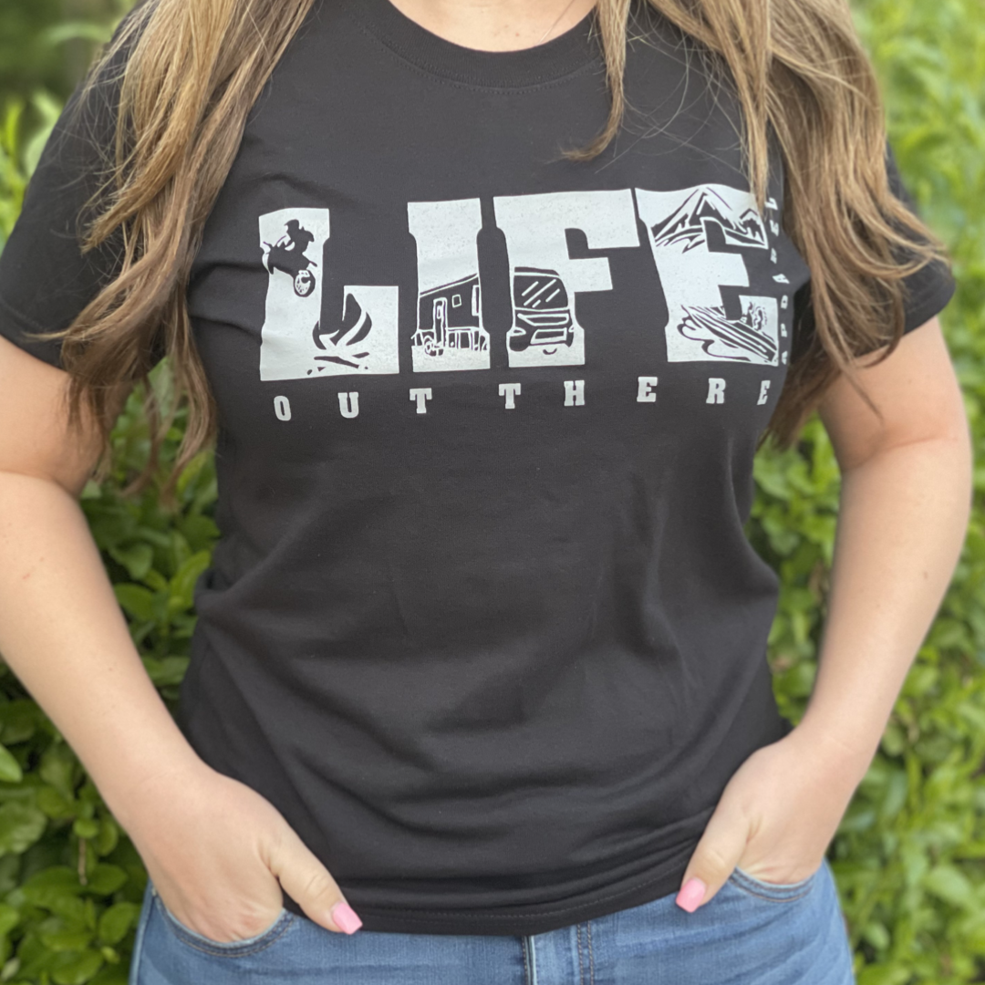A person wearing The OG Women's T Shirt in Black/Grey from Life Out There Apparel, featuring the words LIFE OUT THERE embellished with outdoor-themed illustrations such as camping gear and dirt bikes, stands against a backdrop of green foliage.