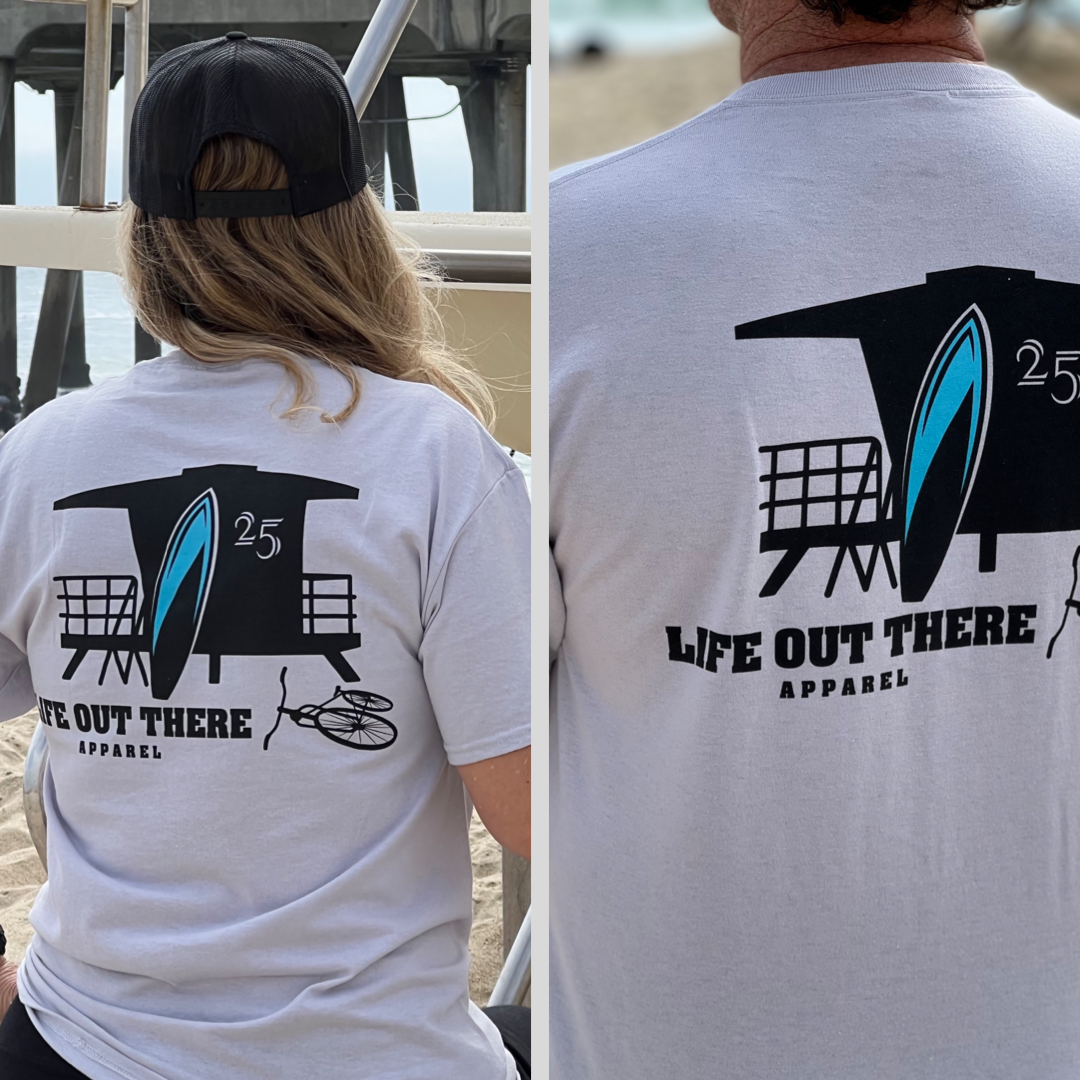 Two individuals are sporting The JJ Tee in silver from Life Out There Apparel, showcasing a design of a lifeguard tower, a surfboard, and the text 25. They are standing with their backs to the camera at Huntington Beach, with sand and the famous pier visible in the background.