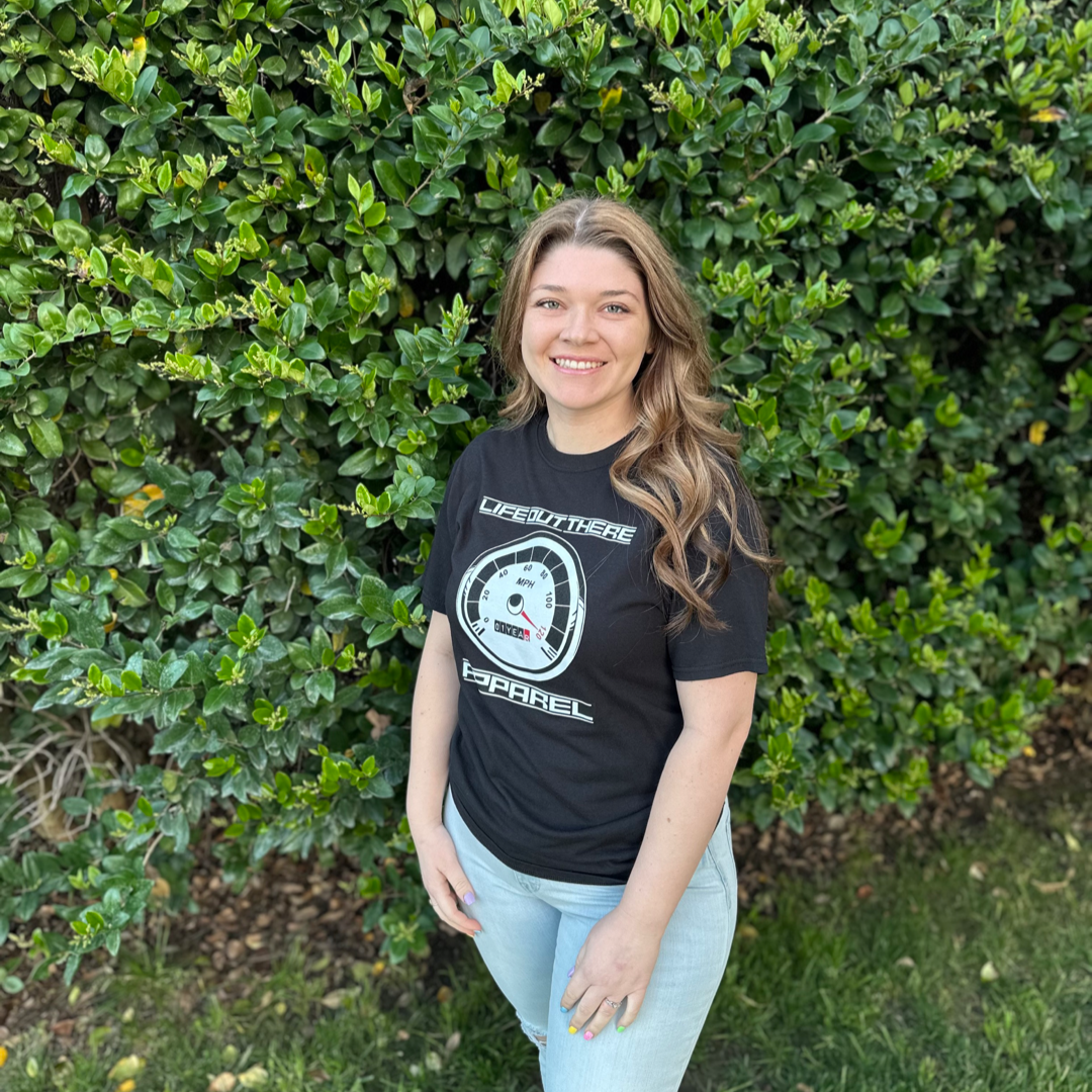 Life Out There Apparel “On My Way” Black Unisex tee shirt is the 1 year anniversary design that has a big speedometer on it - pegged at 120 mph & the odometer is rolling over 1 Year. This photo shows a woman showing the design on the front of the of the shirt standing in front of a wall of green bushes.