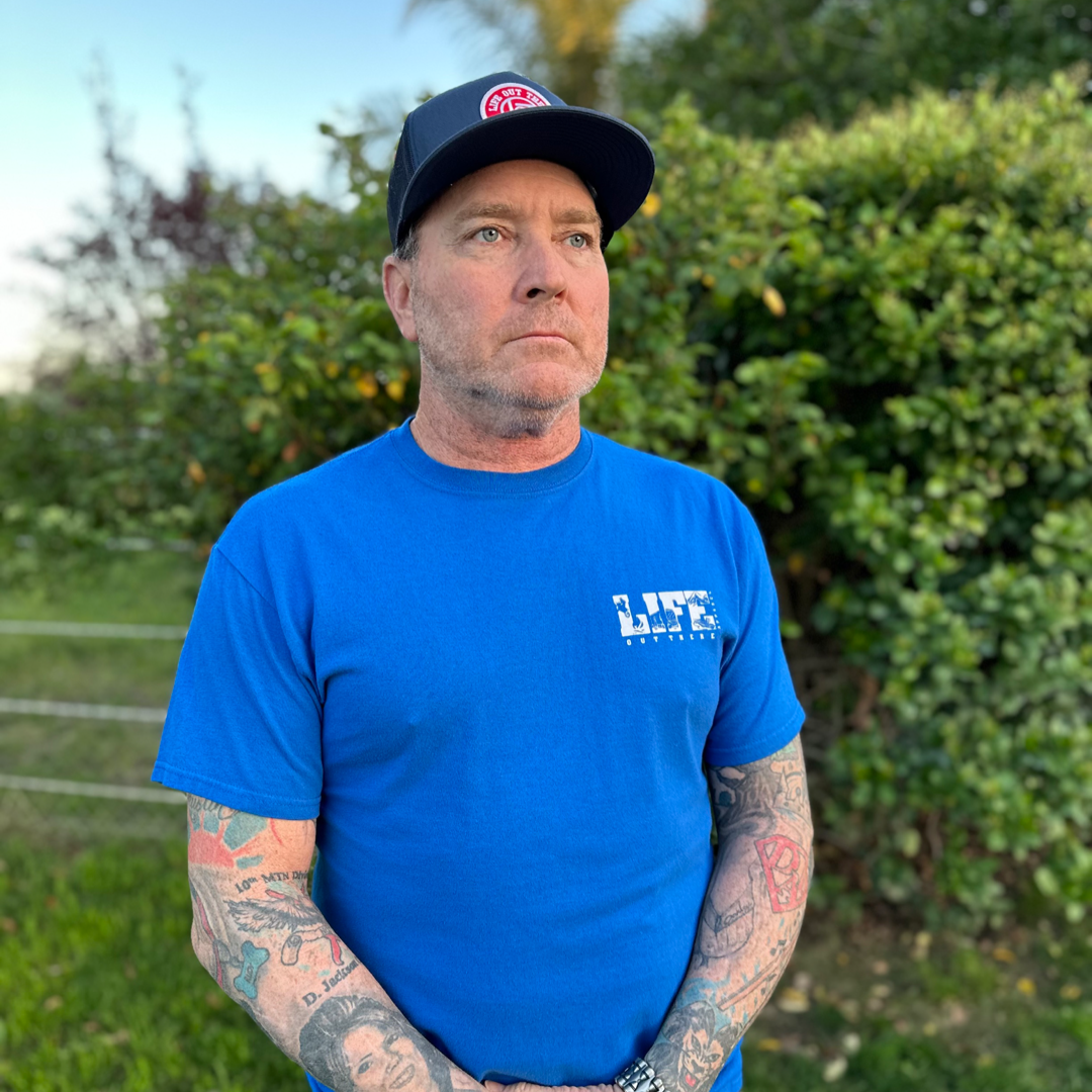 A man with tattooed arms stands outdoors, wearing the Life Out There Apparel "The OG" T-Shirt in royal blue and white and a black cap with a logo. He is in front of lush green foliage and a clear sky, exuding the cool confidence of someone who loves dirt bikes and the thrill of adventure.
