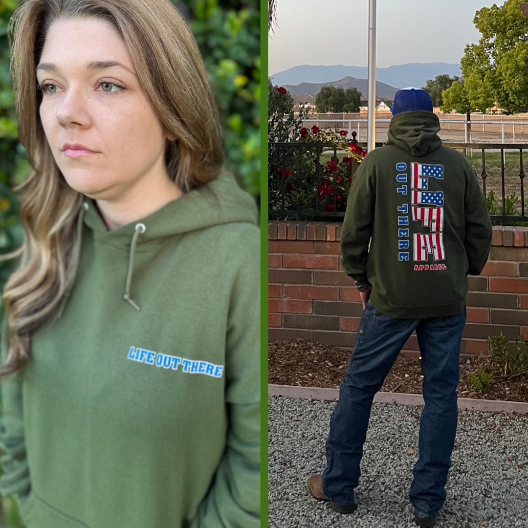 A person dressed in the Unisex The Freedom Hoodie in Military Green by Life Out There Apparel stands outdoors near a garden, with majestic mountains in the backdrop. This hoodie, featuring "Life Out There" on the front and American flags on the back, is perfect for celebrating THE FREEDOM of the 4th of July.