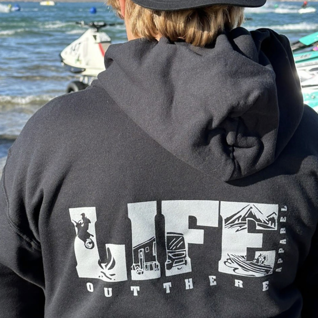 A person wearing the Unisex The OG Hoodie in Black/Grey by Life Out There Apparel stands by a body of water, with jet skis visible in the background. This hoodie embodies outdoor activities such as motorcycling, RV camping, and snowboarding, perfectly capturing the adventurous spirit of life beyond the ordinary.