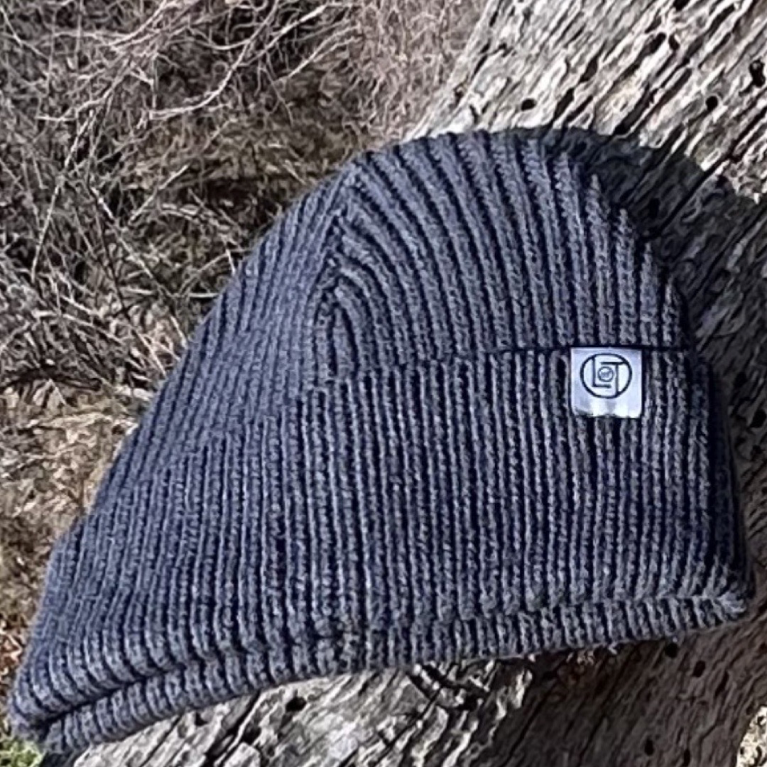 The Men's Knit Beanie - Charcoal from Life Out There Apparel, featuring a small white logo patch, rests elegantly on a tree stump with dry, brown foliage in the background.
