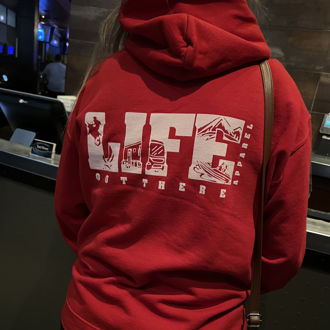 A person is wearing the Unisex The OG Hoodie in red and white by Life Out There Apparel, featuring outdoor-themed graphics on the back. They're standing at a counter in a dimly lit setting, capturing the adventurous spirit of snowboarding under starry winter skies.