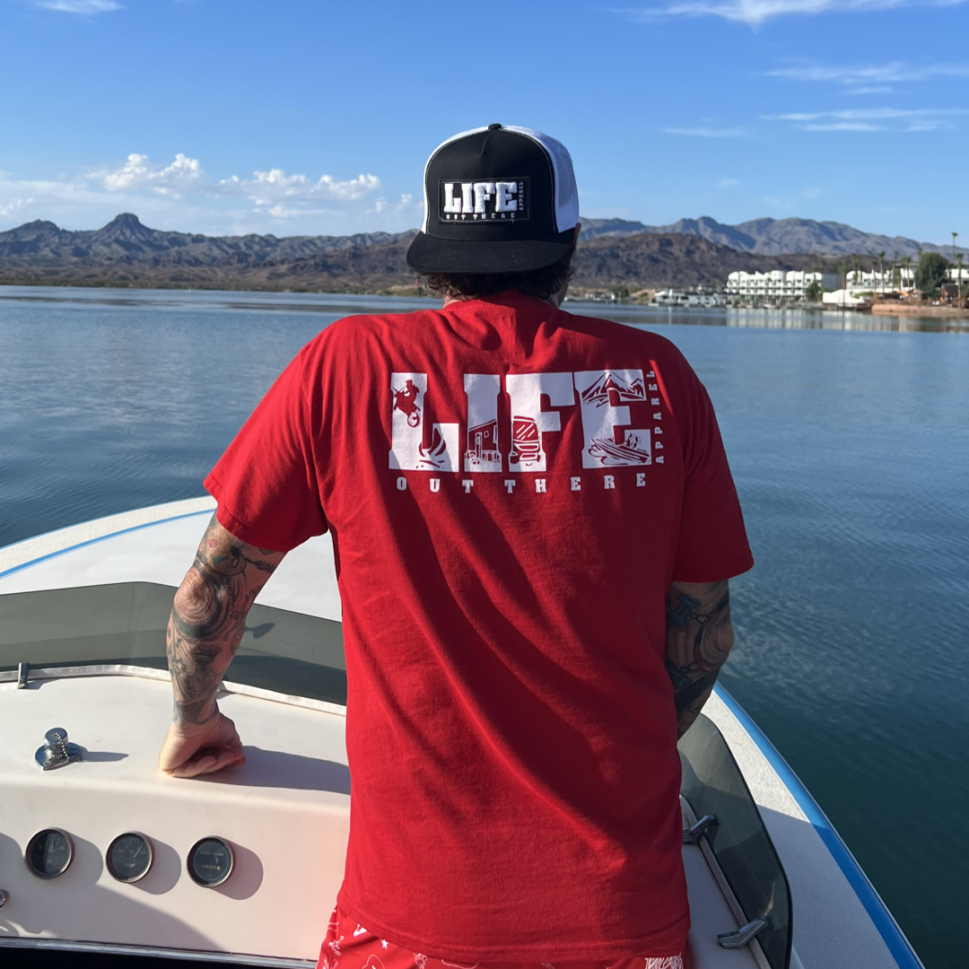 A person sporting the Unisex The OG T Shirt in red and white from Life Out There Apparel, along with a black-and-white cap, stands at the front of a boat, gazing over a tranquil lake. In the background, mountains reminiscent of those ideal for snowboarding rise under a blue sky scattered with distant buildings.