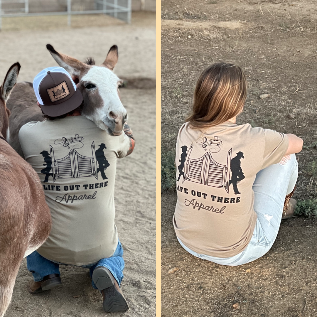 Left: A person wearing a sandstone-colored Unisex THE YEEHAW T Shirt and cap embraces a donkey in a sandy area. Right: Another individual, also sporting the same cowboy-themed shirt from Life Out There Apparel with text printed on the back, sits on grass facing away, embodying the true spirit of YEEHAW.