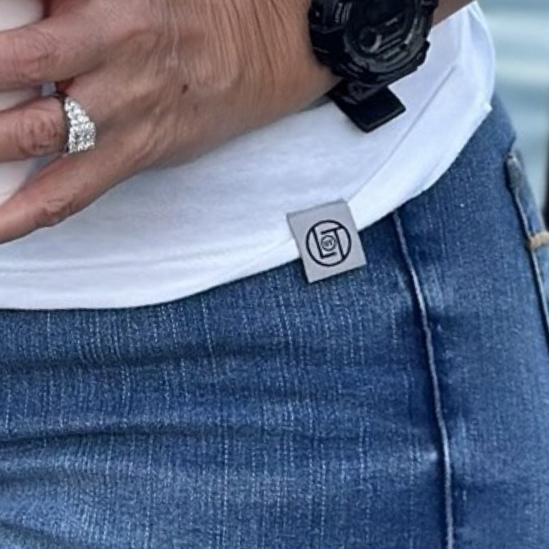 A close-up of someone dressed in The Havasu Women's T Shirt - White from Life Out There Apparel, paired with blue jeans for a summer vibe. They have a ring on their finger and are wearing a black watch, and a small logo tag on the shirt accentuates the relaxed elegance so often associated with the Lake Havasu area.