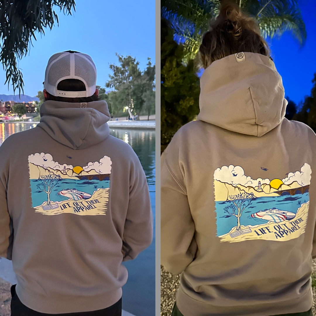 Two individuals donning the Unisex The Havasu Hoodie - Rock by Life Out There Apparel, featuring a vibrant graphic of a coastal scene and the phrase "Life out here," are standing outdoors amidst water and trees at Windsor Beach. One enjoys the daylight, while the other is surrounded by dusk's glow, embodying Lake Havasu's tranquil allure.