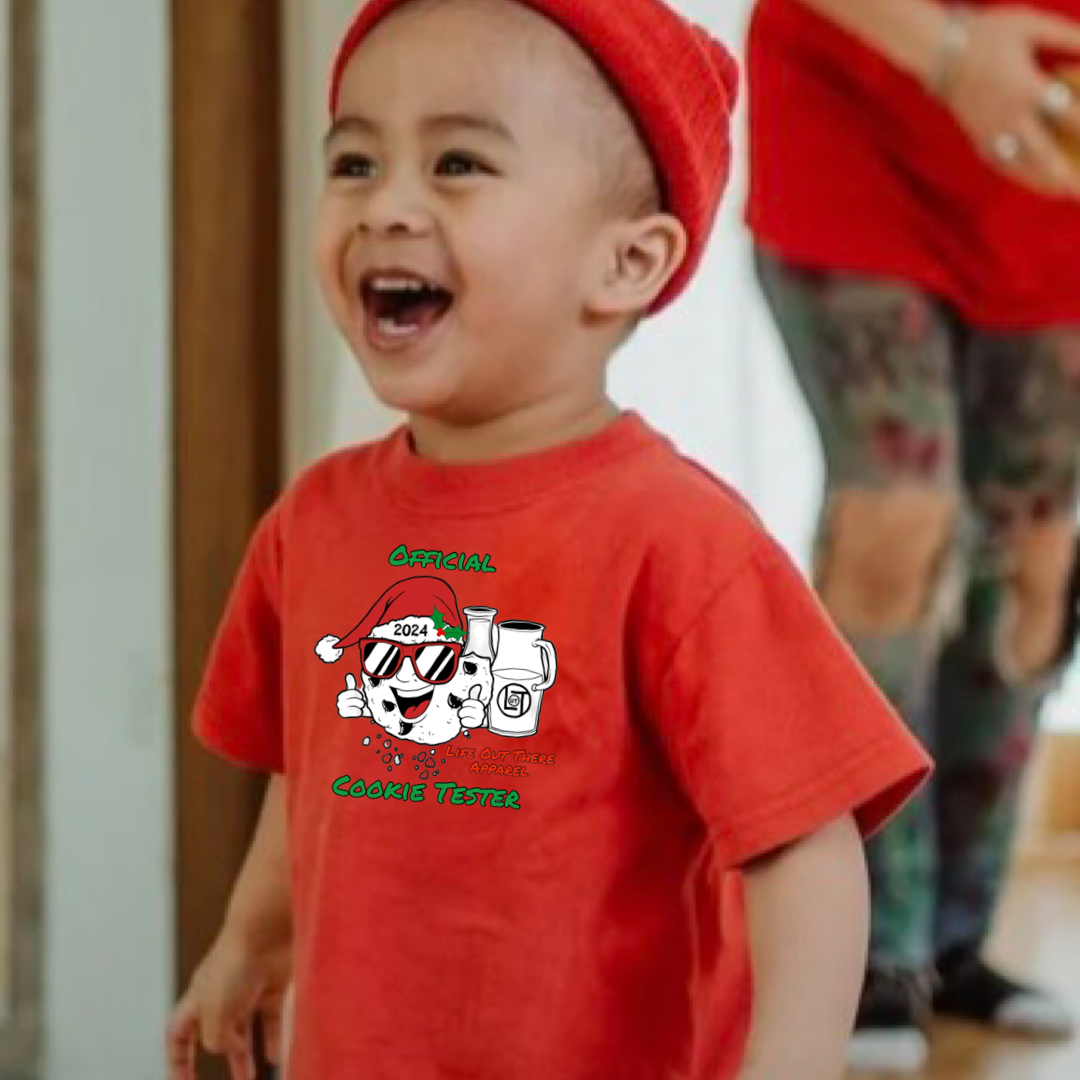 Red short sleeve toddler t-shirt with a cartoon cookie wearing a Santa hat and sunglasses labeled "2024," alongside glass milk cartons. Text reads, "Life Out The Apparel / official cookie tester”.