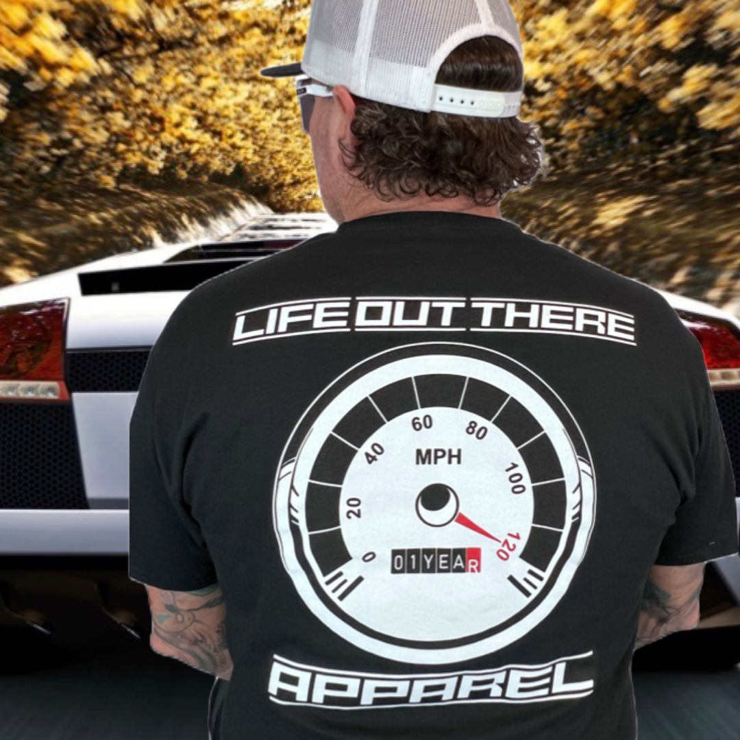 Life Out There Apparel “On My Way” Black Unisex tee shirt is the 1 year anniversary design that has a big speedometer on it - pegged at 120 mph & the odometer is rolling over 1 Year. This photo shows a man showing the design on the back of the shirt standing behind a white sports car.