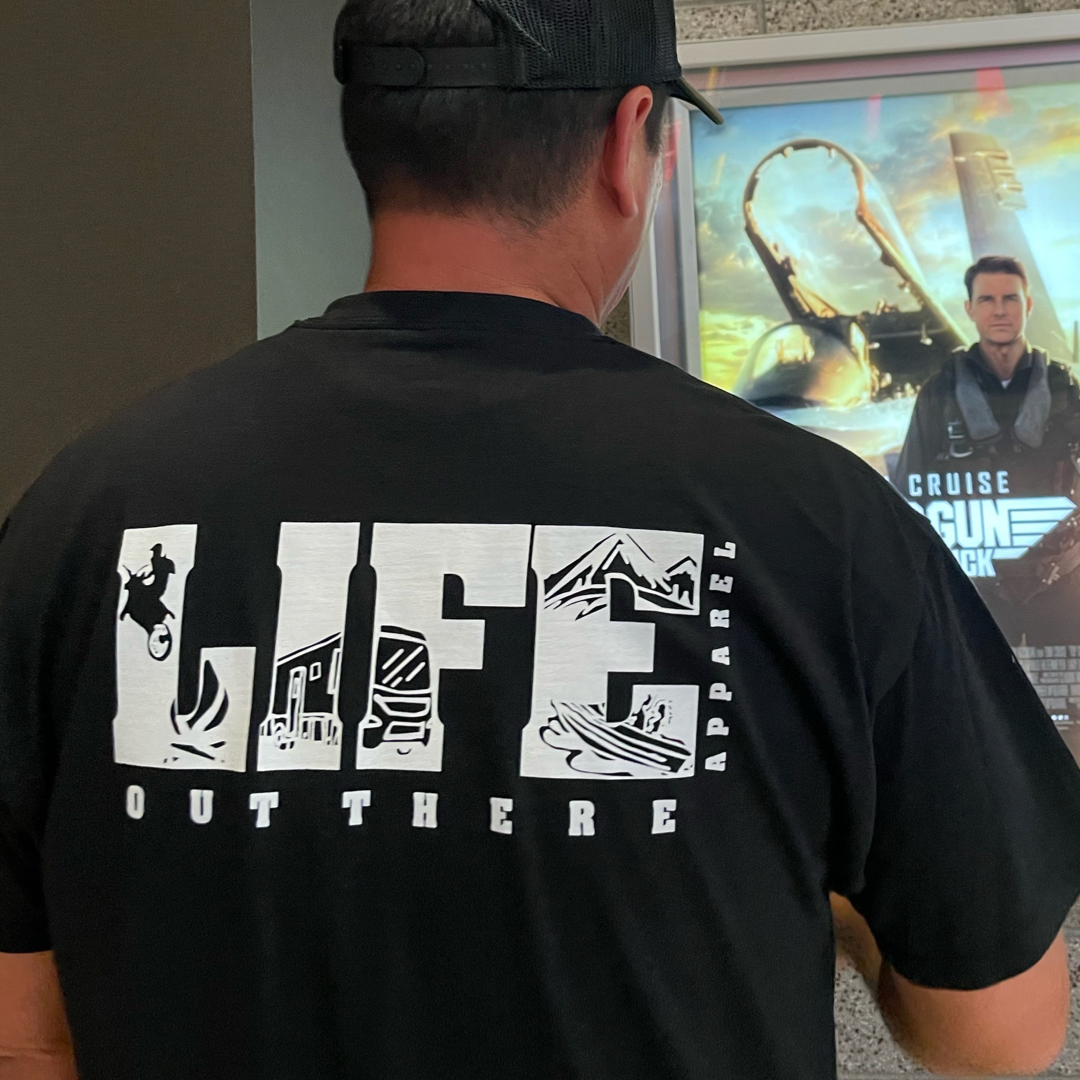 A person wearing the Unisex The OG T Shirt in Black/White from Life Out There Apparel stands in front of a movie poster. The shirt, featuring outdoor-themed designs, evokes adventures such as snowboarding under starry skies or camping by rugged trails.