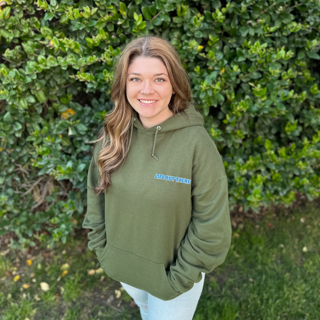 A person with long hair stands outdoors, smiling against a backdrop of green leaves. They're wearing the Unisex The Freedom Hoodie in Military Green by Life Out There Apparel and light jeans, embodying the American spirit of freedom.