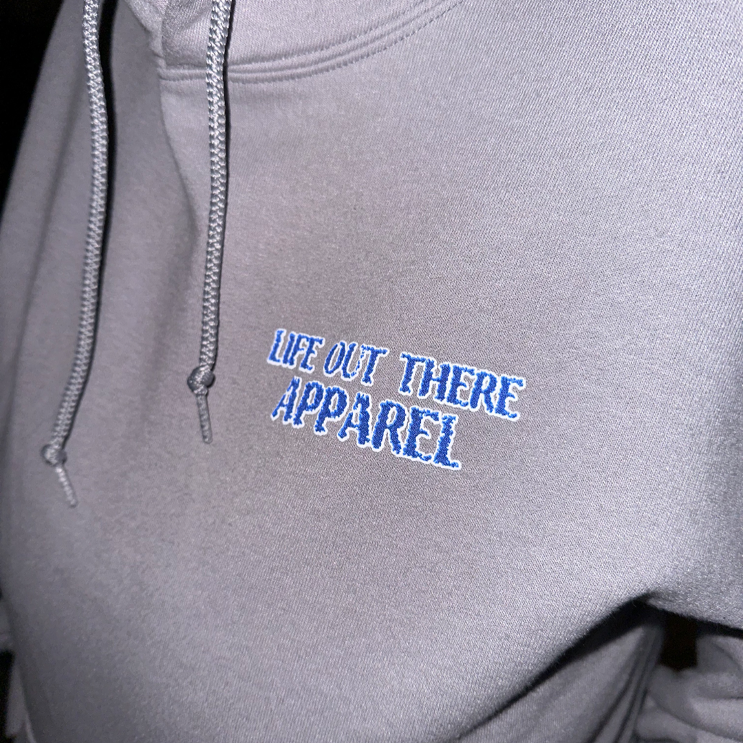A close-up of the Unisex The Havasu Hoodie - Rock showcases its sleek gray design with Life Out There Apparel embroidered in blue on the chest, making it perfect for breezy days at Windsor Beach. The hoodie includes drawstrings near the neckline.