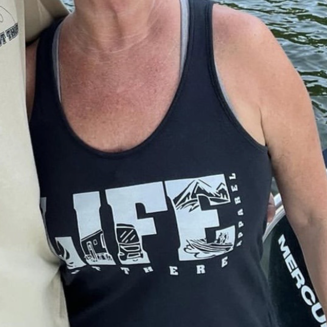 A person is wearing The OG Women's Tank Top in black and grey by Life Out There Apparel, featuring outdoor-themed graphics. The background displays greenery similar to a camping trip, with another person's arm partly visible.