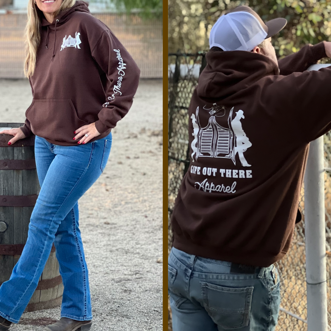 Two individuals wearing the Unisex THE YEEHAW Hoodie in Chocolate/White from Life Out There Apparel embody ranch life. The first person leans on a barrel, highlighting the front design, while the second stands with their back to a chain-link fence and trees, perfectly capturing that YEEHAW spirit.