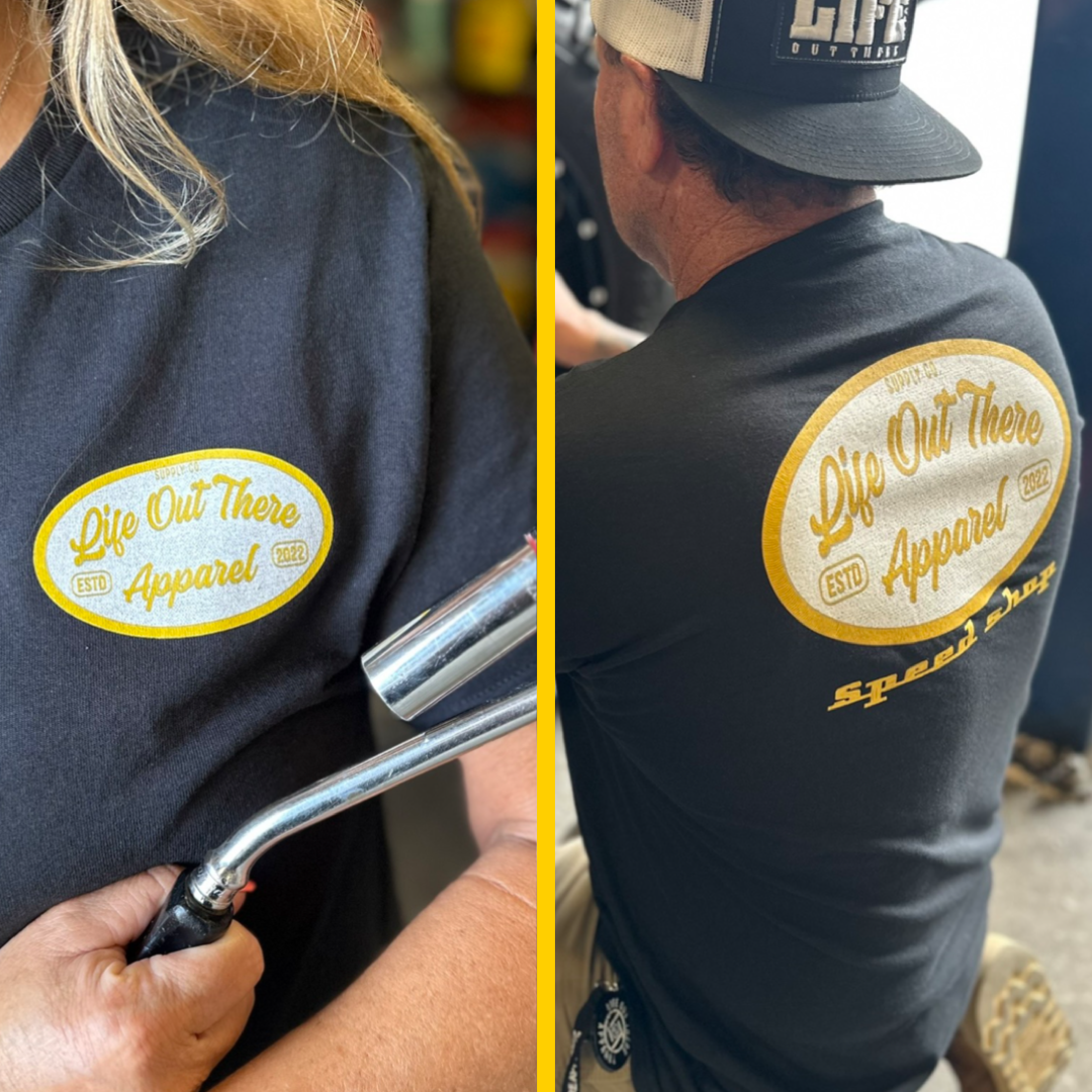 Two individuals are sporting the Unisex Speed Shop T-shirt in black featuring yellow oval logos that read "Life Out There Apparel ESTD 2022." The back of one shirt displays the phrase "speed surf" beneath the logo. One person holds a tool reminiscent of car customization, and both wear caps decorated with embroidered patches.
