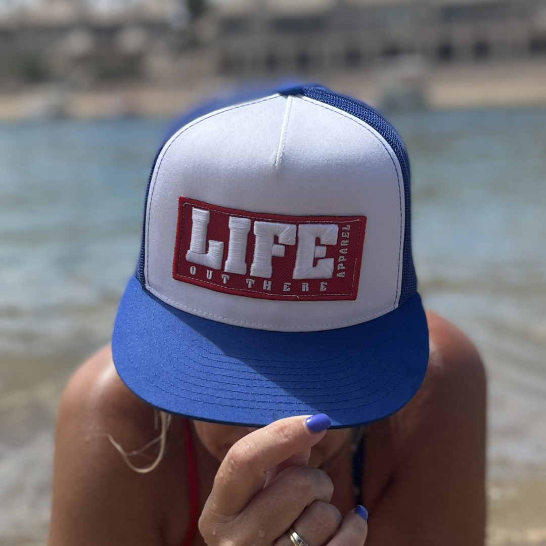 A person wearing a Royal/White Life Out There - Puff Embroidered SnapBack Trucker Hat from Life Out There Apparel is crouching by the water, with a blurred background. Their nails are painted blue.