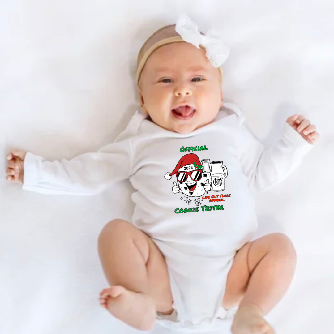 White long sleeve baby shirt with a cartoon cookie wearing a Santa hat and sunglasses labeled "2024," alongside glass milk cartons. Text reads, "Life Out The Apparel / official cookie tester”.