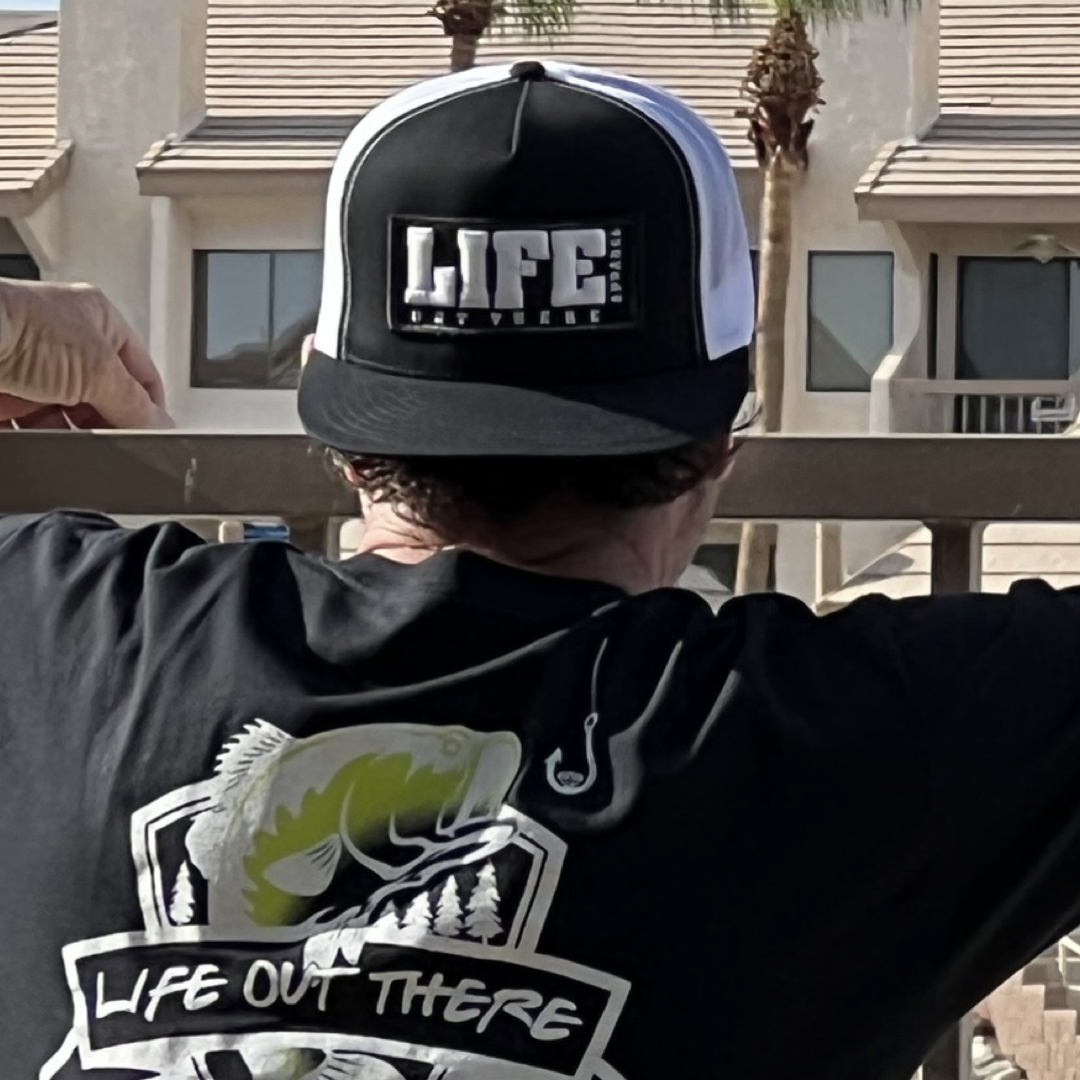 A person sporting a Life Out There - Puff Embroidered SnapBack Trucker Hat in black and white, along with a black t-shirt featuring a fish graphic and the phrase "Life Out There," leans against a balcony railing, facing away. The background showcases palm trees and a building with large windows.