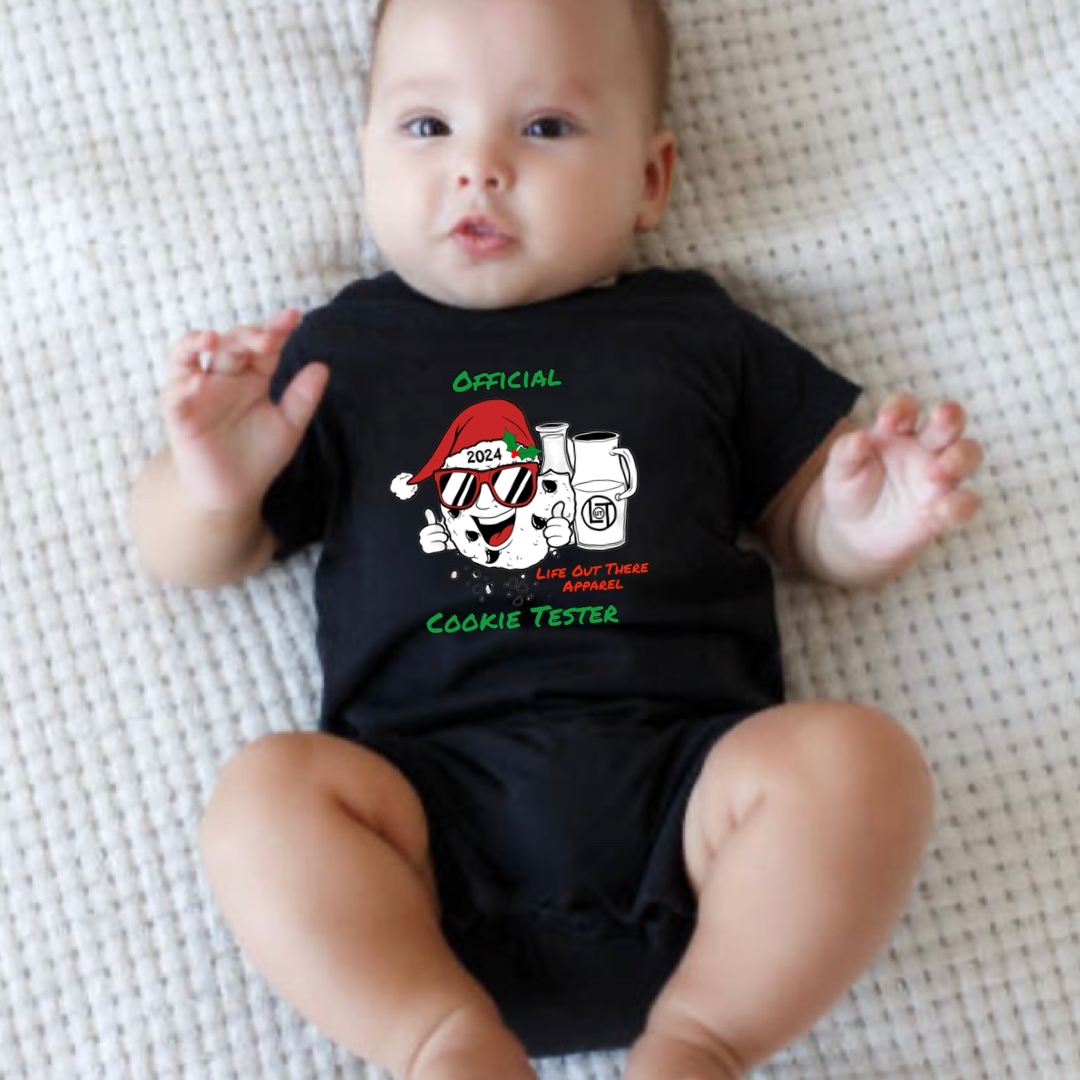 Black short sleeve baby shirt with a cartoon cookie wearing a Santa hat and sunglasses labeled "2024," alongside glass milk cartons. Text reads, "Life Out The Apparel / official cookie tester”.