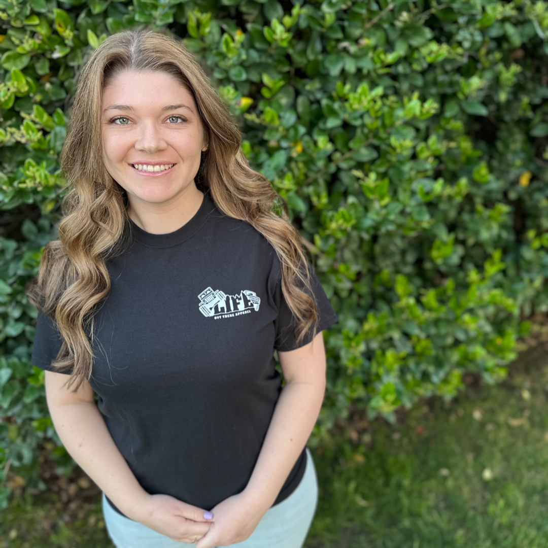 A person with long wavy hair is standing outdoors in front of green foliage, smiling and wearing a black "Unisex The Crawler T Shirt" from Life Out There Apparel.