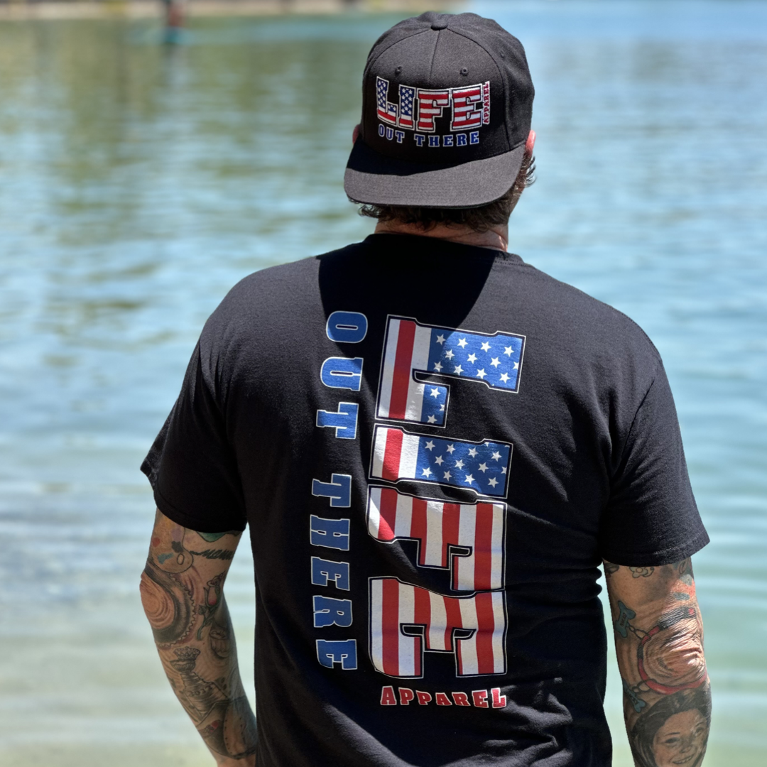 A person with tattoos stands by a body of water, wearing a black hat and the "Unisex The Freedom T Shirt - Black" by Life Out There Apparel, featuring "USA Out There" with stars and stripes design elements, embodying the spirit of freedom in their patriotic apparel.
