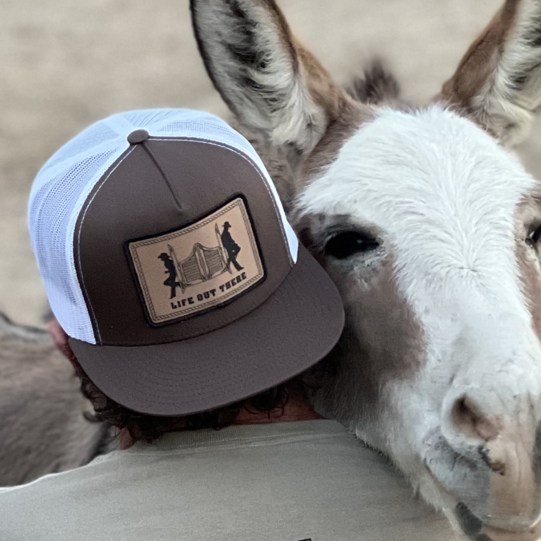 A person wearing the Life Out There - THE YEEHAW SnapBack Trucker Hat in brown and white from Life Out There Apparel is embracing a donkey. The donkey's head rests gently on the person's shoulder, capturing an affectionate moment that underscores the timeless appeal of this classic trucker style.