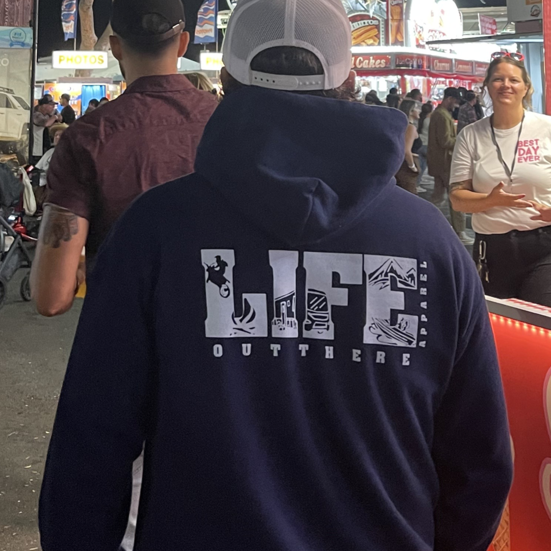 A person wearing a Unisex The OG Hoodie in Navy/White from Life Out There Apparel strolls through a bustling outdoor fair, reminiscent of camping adventures. Stalls with neon lights and lively crowds fill the background, and a banner overhead adds to the festive atmosphere.