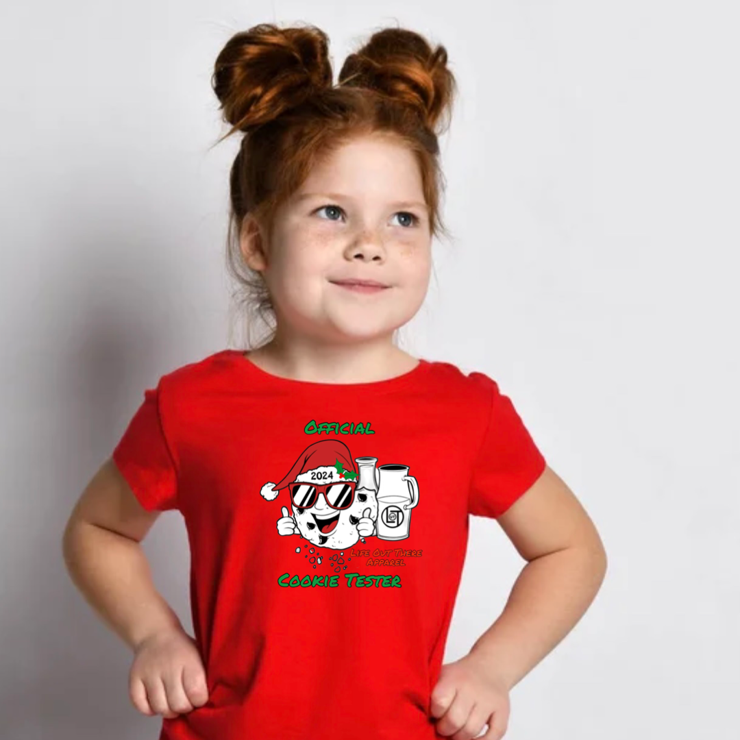 Red short sleeve toddler t-shirt with a cartoon cookie wearing a Santa hat and sunglasses labeled "2024," alongside glass milk cartons. Text reads, "Life Out The Apparel / official cookie tester”.