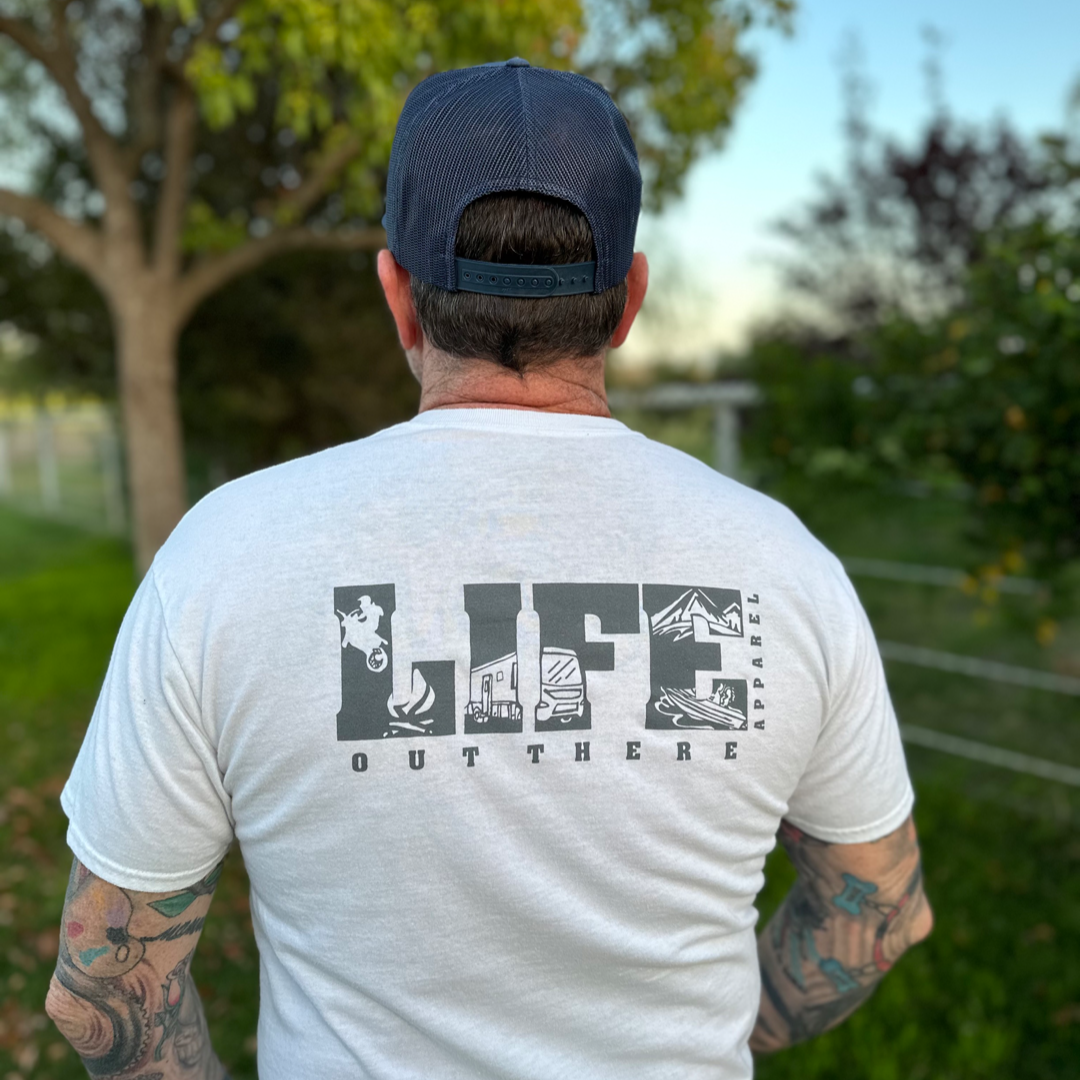 A person stands outdoors, wearing the Unisex The OG T-Shirt in white and grey from Life Out There Apparel, featuring nature-themed illustrations. They have tattoos on their arms and are sporting a blue cap, giving off an adventurous vibe perfect for dirt biking. The setting includes trees and a grassy area in the background.