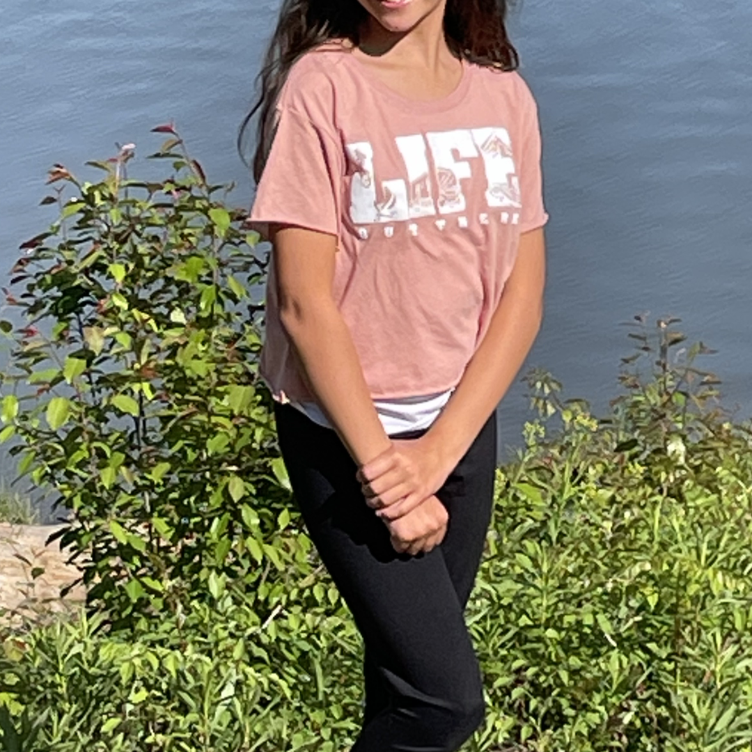 A girl wearing the "OG Women's Crop T-Shirt" in Desert Pink/White by Life Out There Apparel and black pants stands near a lush green bush by a body of water, dreaming of her next camping adventure. The sun is shining, casting a warm glow on her long brown hair.