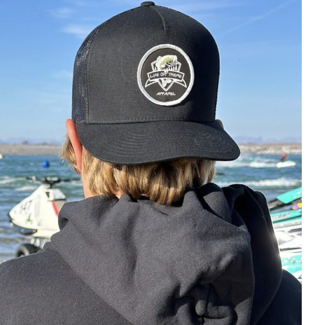 A person in a black hoodie and The Hooker Snapback Trucker Hat by Life Out There Apparel faces a scenic lake dotted with jet skis. The cap, featuring a fish logo, enhances the serene view of a clear sky sprinkled with clouds.
