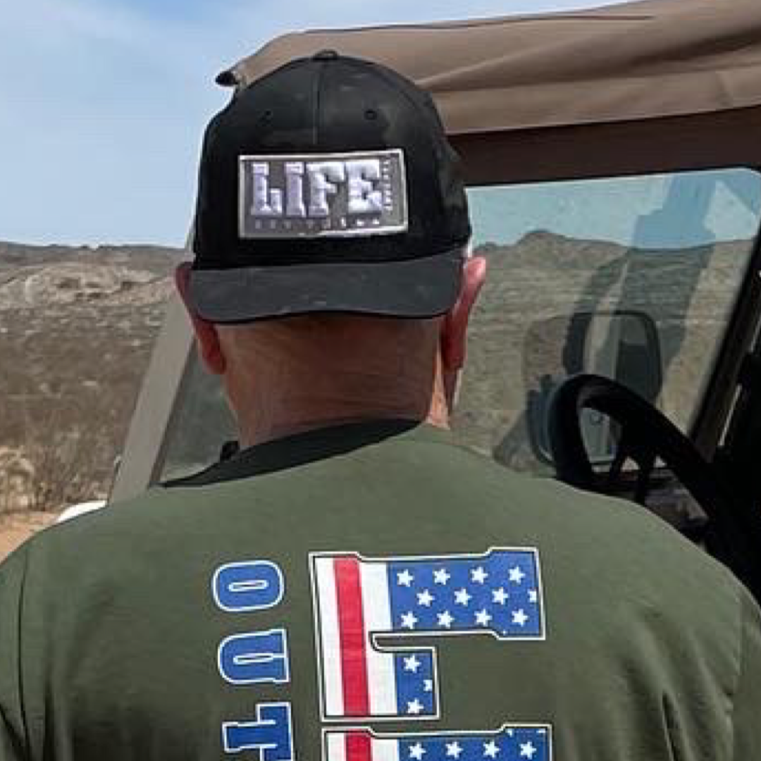 A person wearing a black camo SnapBack hat from Life Out There Apparel, featuring puff embroidery, with the word LIFE stands next to a Jeep in a desert landscape. They are also wearing a green shirt showcasing the word OUTDOORS and an American flag design. Hills and a clear sky are visible in the background.