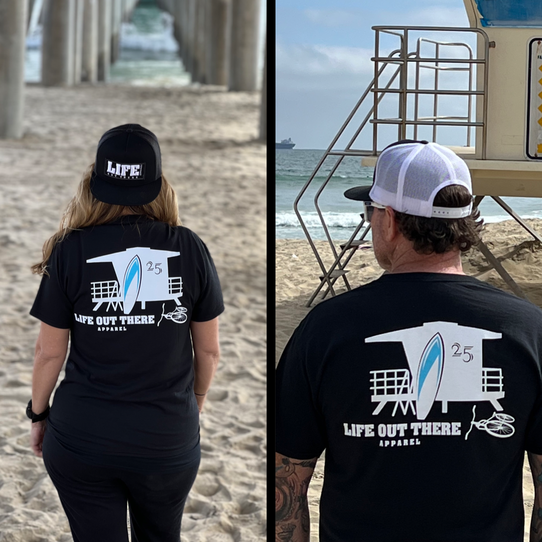 A couple, both wearing the Unisex The JJ T Shirt in black by Life Out There Apparel, enjoy their summer getaway. One of them complements their outfit with a LIFE text cap. They're standing on a sandy beach, with the ocean and a pier creating an idyllic scene in the background.