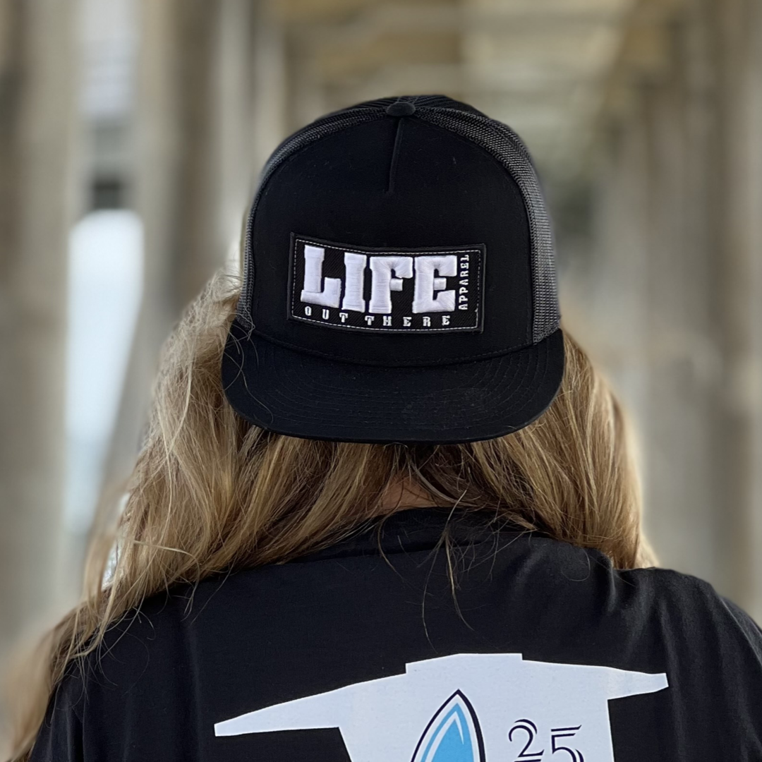 A person with long hair is wearing the Life Out There - Puff Embroidered SnapBack Trucker Hat in black from Life Out There Apparel, standing under a pier with blurred columns visible in the background.