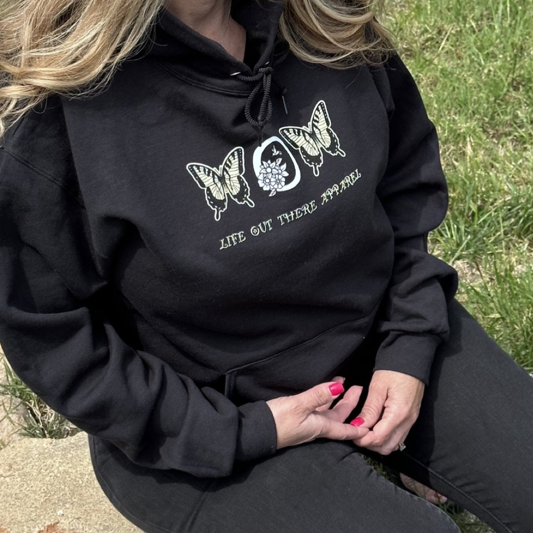 A person wearing the Love Mom Hoodie in black from Life Out There Apparel sits on the grass outdoors. They're dressed in dark pants and have pink nail polish, creating a serene scene that evokes a Mother's Day card.