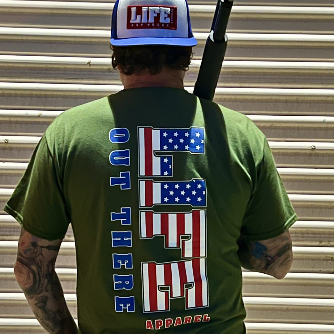 Dressed in a military green "The Freedom T-Shirt" by Life Out There Apparel, featuring American flag-themed lettering with an "Out There" design, a person stands proudly before a corrugated metal wall. Celebrating the 4th of July spirit, they wear a blue and white cap with "LIFE" on it, embodying the Land of the Free while holding an object over their shoulder.