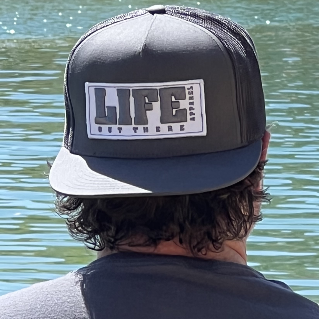 A man with curly hair, wearing a grey SnapBack trucker hat by Life Out There Apparel, sits by a serene body of water. The calm surface perfectly mirrors the sunlight as he gazes into the distance.