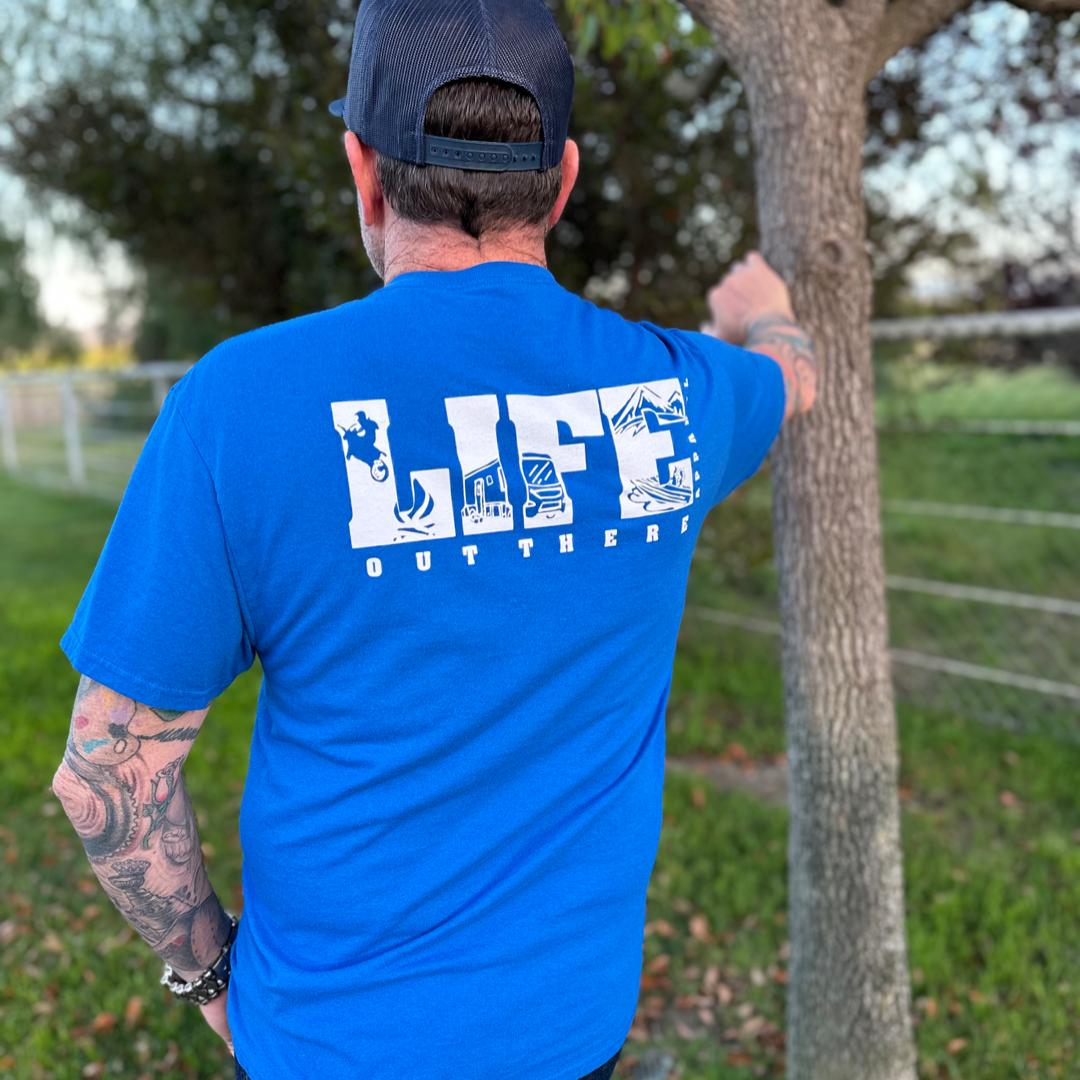 A man wearing a blue Unisex The OG T Shirt in Royal/White by Life Out There Apparel and a black cap leans against a tree, with tattoos visible on his arms. The grassy area with trees and fencing suggests an ideal spot for camping adventures.