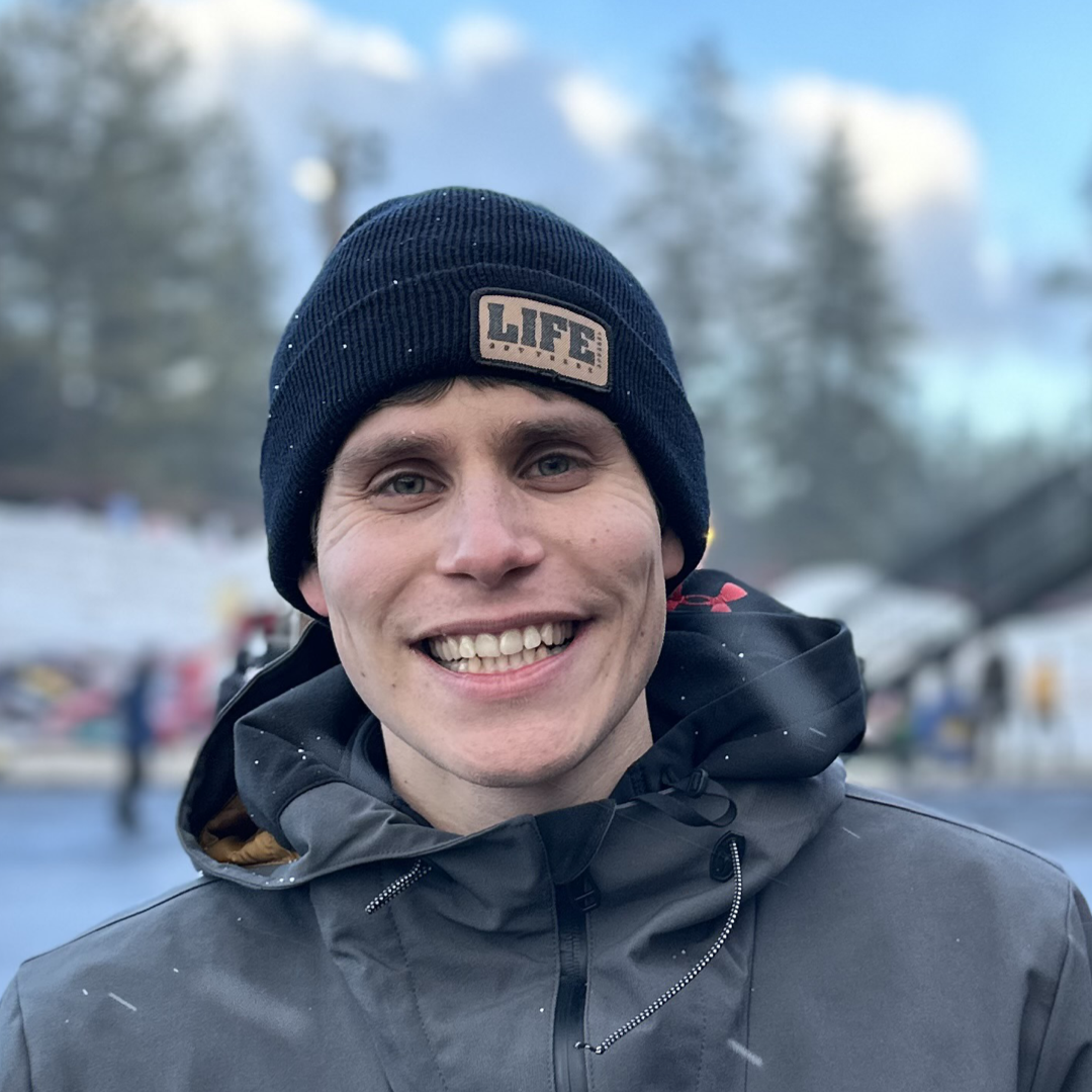 A person smiling outdoors, wearing a Life Out There - Cuff Beanie in black by Life Out There Apparel and a gray jacket. The background is slightly blurry with trees and a snowy landscape.
