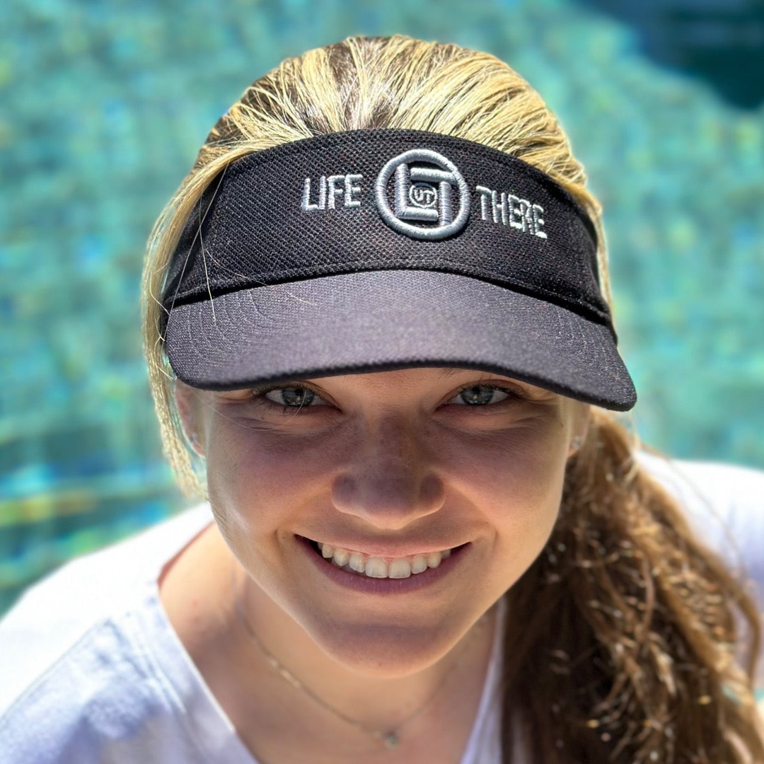 A person smiling, wearing a black visor from Life Out There Apparel's Summer Embroidered Sun Visors collection, standing in front of a blurred blue-tinted background.