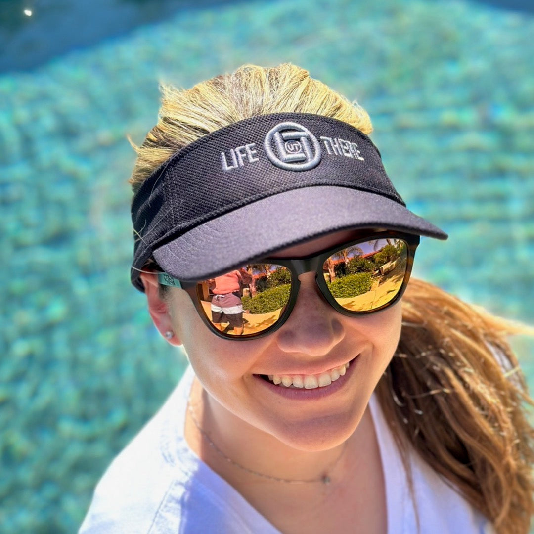 A woman stands by a pool, smiling and wearing sunglasses along with a Life Out There - Summer Embroidered Sun Visor from Life Out There Apparel. The water reflects in her sunglasses under the bright and clear sky.