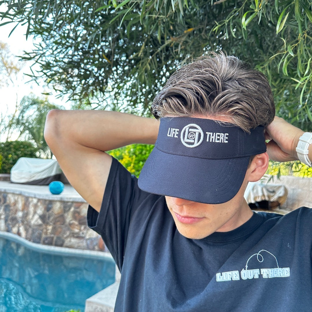 A person wearing a Life Out There Apparel - Summer Embroidered Sun Visor and a matching T-shirt with "Life Out There" printed on it is standing by a pool. With their hands behind their head, the scene is set against a backdrop of trees and a poolside area.